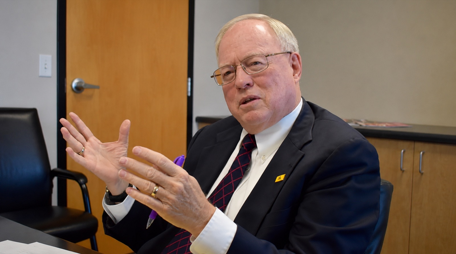 Hans Andersen in a dark suit and red tie, seated at a conference table mid-conversation, gesturing as he explains a point.