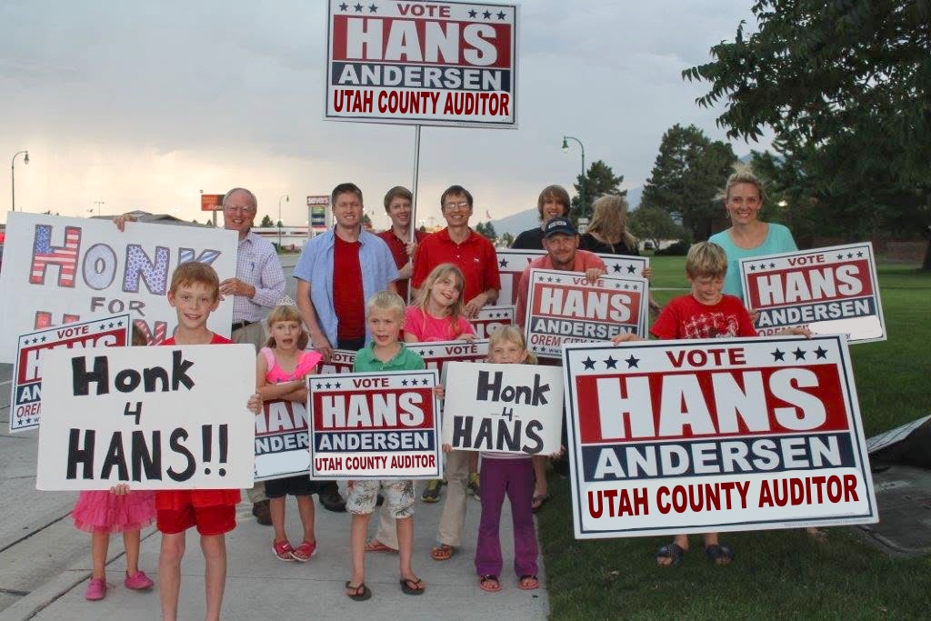 A roadside crowd of supporters — families and children — holding hand-lettered "Honk 4 Hans" signs alongside printed "Vote Hans Andersen — Utah County Auditor" campaign signs at dusk.