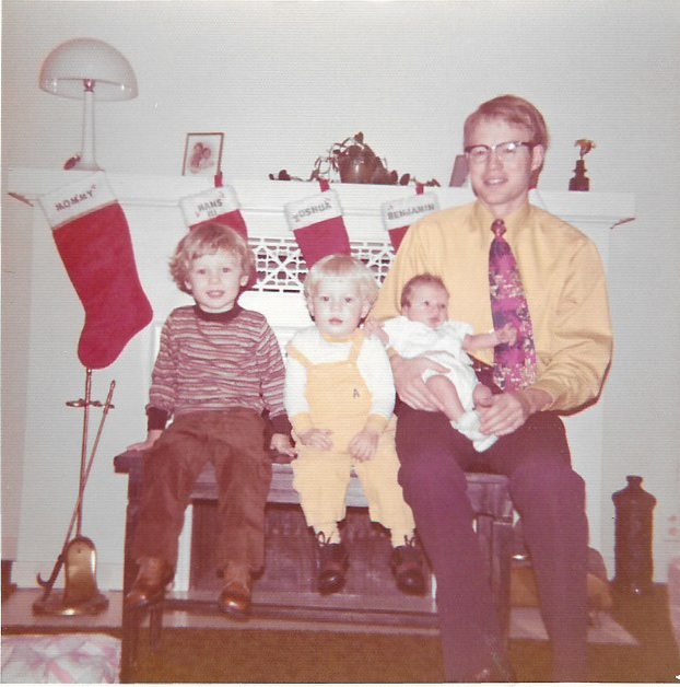 A 1970s family photo: a young Hans Andersen in a yellow shirt and tie sits on the hearth holding his infant son, with two of his small boys beside him and Christmas stockings hung on the mantel.