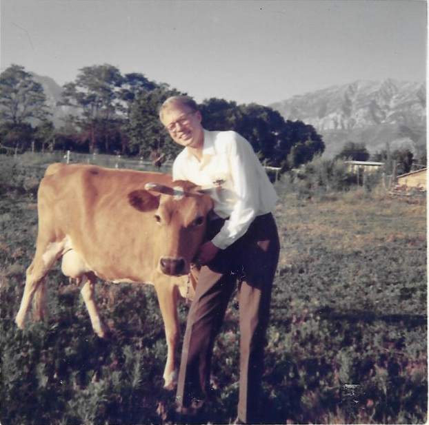 A young Hans Andersen in a white shirt and slacks, smiling as he stands beside a jersey cow in a green pasture, with the Wasatch mountains behind him.