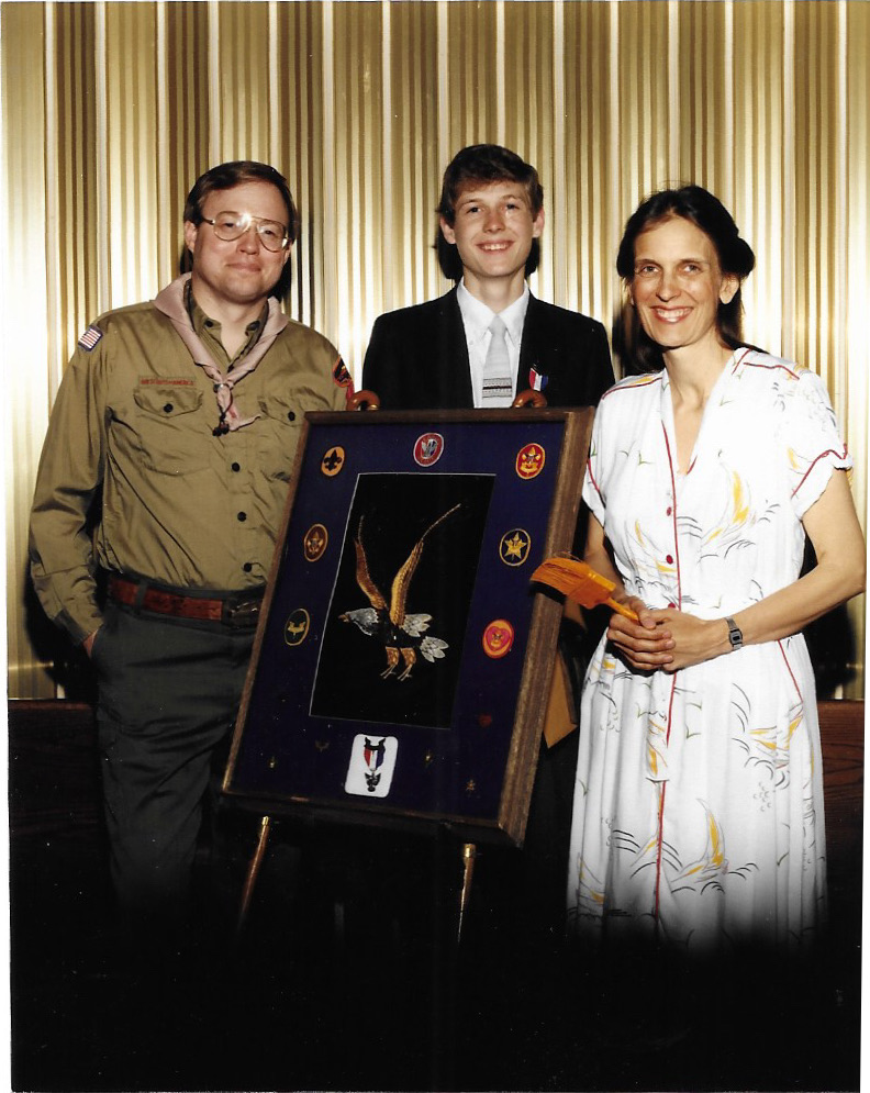 Hans Andersen in his Scoutmaster uniform at an Eagle Scout court of honor, standing with the new Eagle Scout and his family beside a framed display of merit badges and the Eagle emblem.